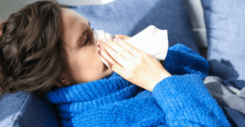 sick breastfeeding mother laying down on blue sofa wearing a blue cozy jacket and blowing her nose